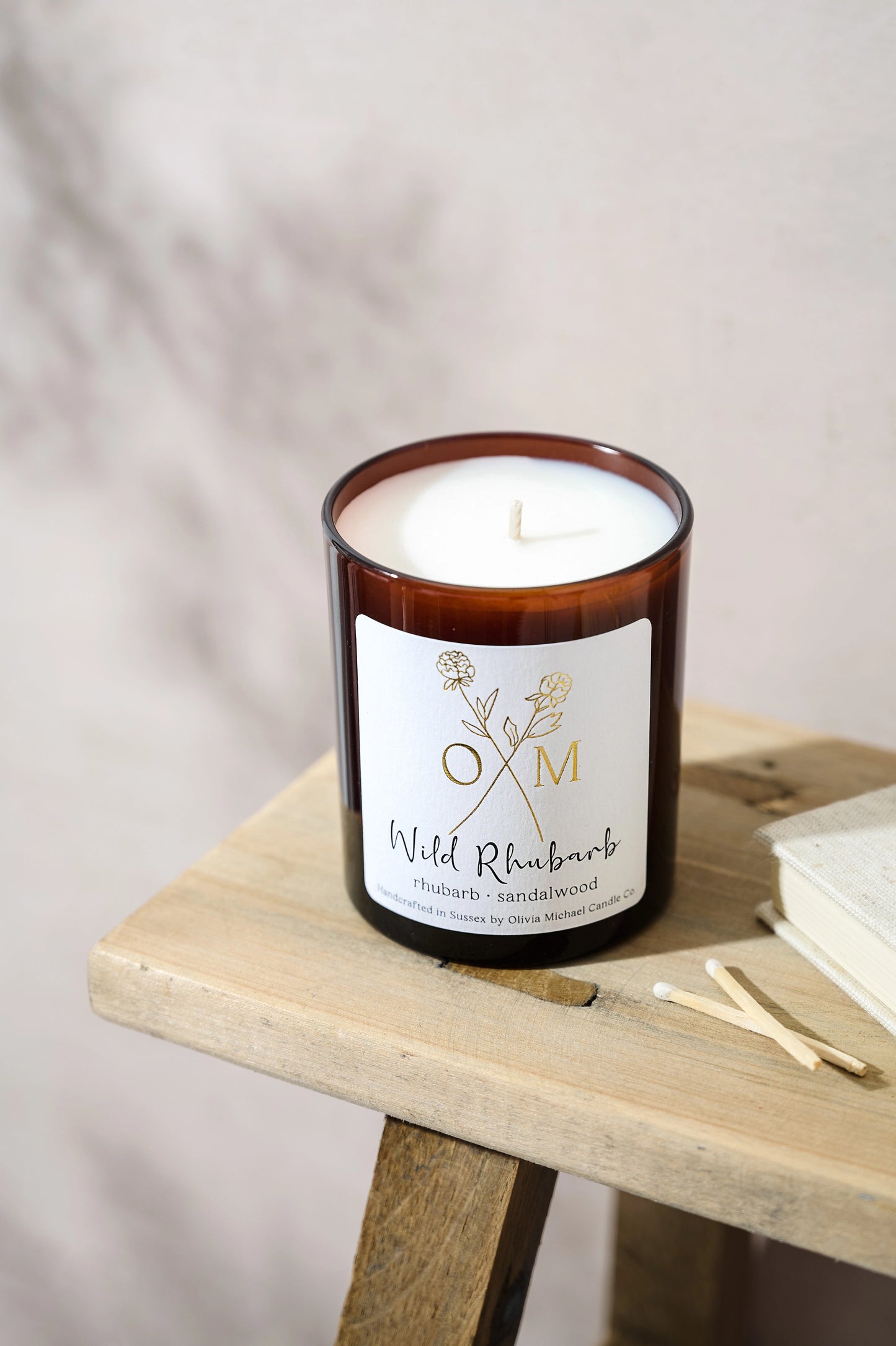 Wild Rhubarb scented candle in a brown glass jar with an unlit white wax wick, placed on a wooden surface next to a book and two matches. The label reads 'Wild Rhubarb, rhubarb and sandalwood'.