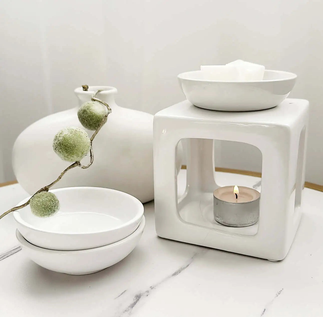 Minimalist wax melt burner set with a lit tealight candle, white ceramic bowls, and a decorative frosted greenery stem on a white marble surface.