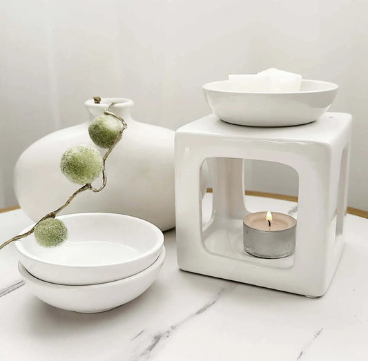 Minimalist wax melt burner set with a lit tealight candle, white ceramic bowls, and a decorative frosted greenery stem on a white marble surface.