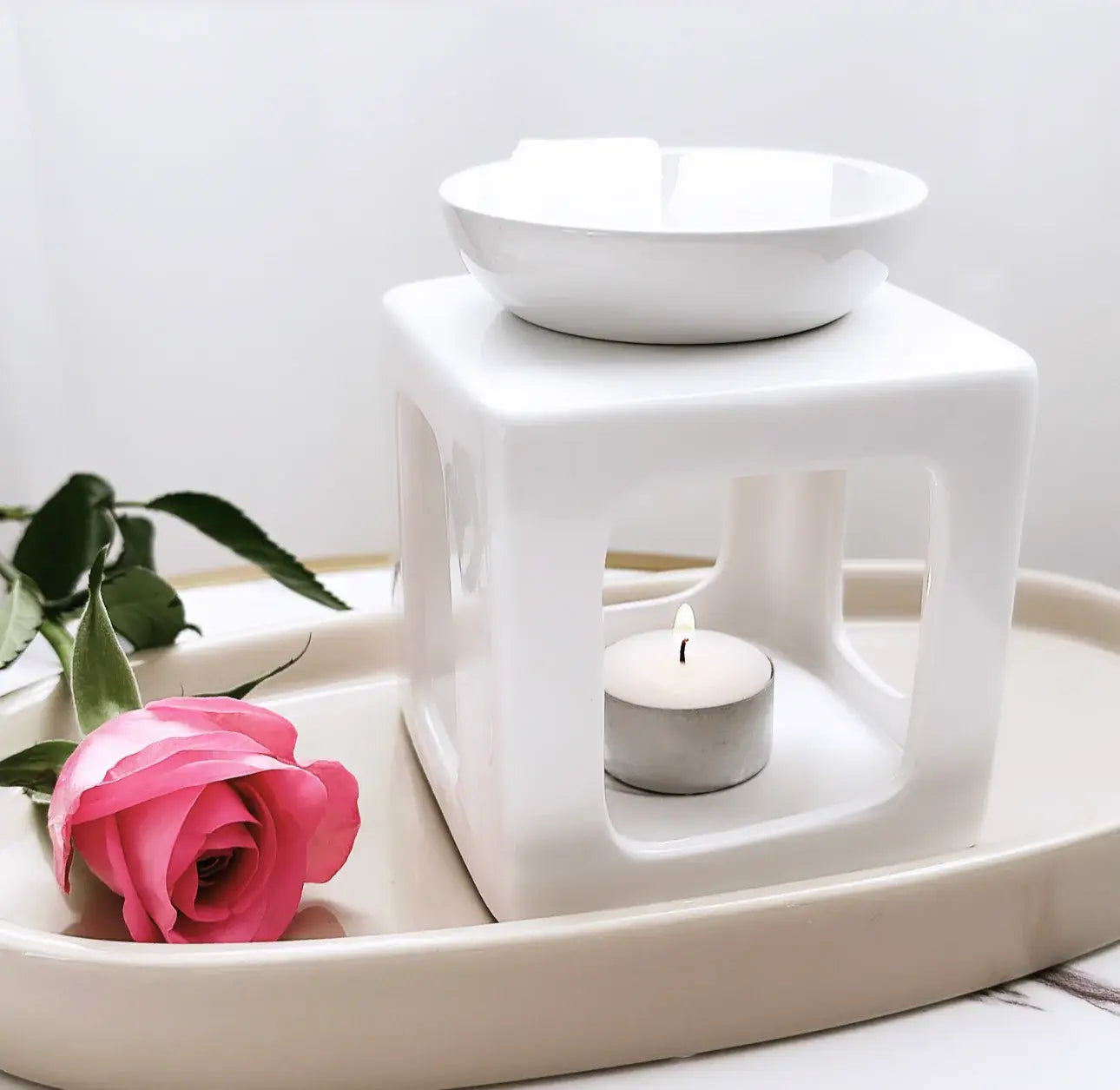 Elegant white ceramic wax melt burner with a lit tealight candle, placed on a cream tray beside a fresh pink rose.