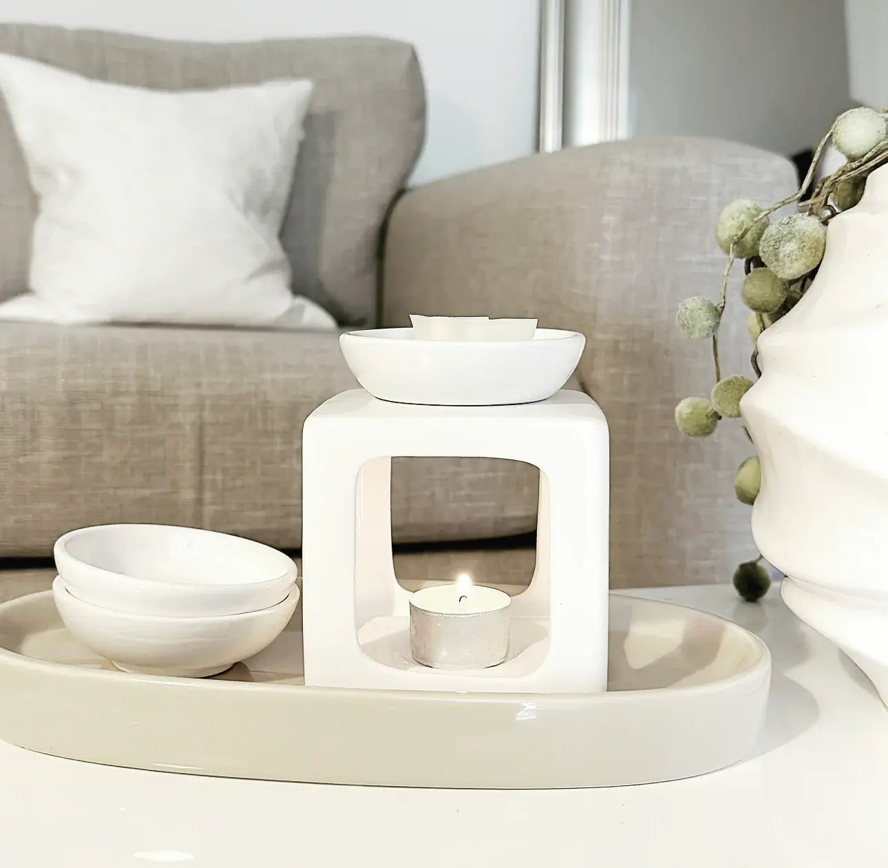 Stylish ceramic wax melt burner with a lit tealight candle on a neutral tray, set in a cozy living room with a beige sofa and decorative accents.