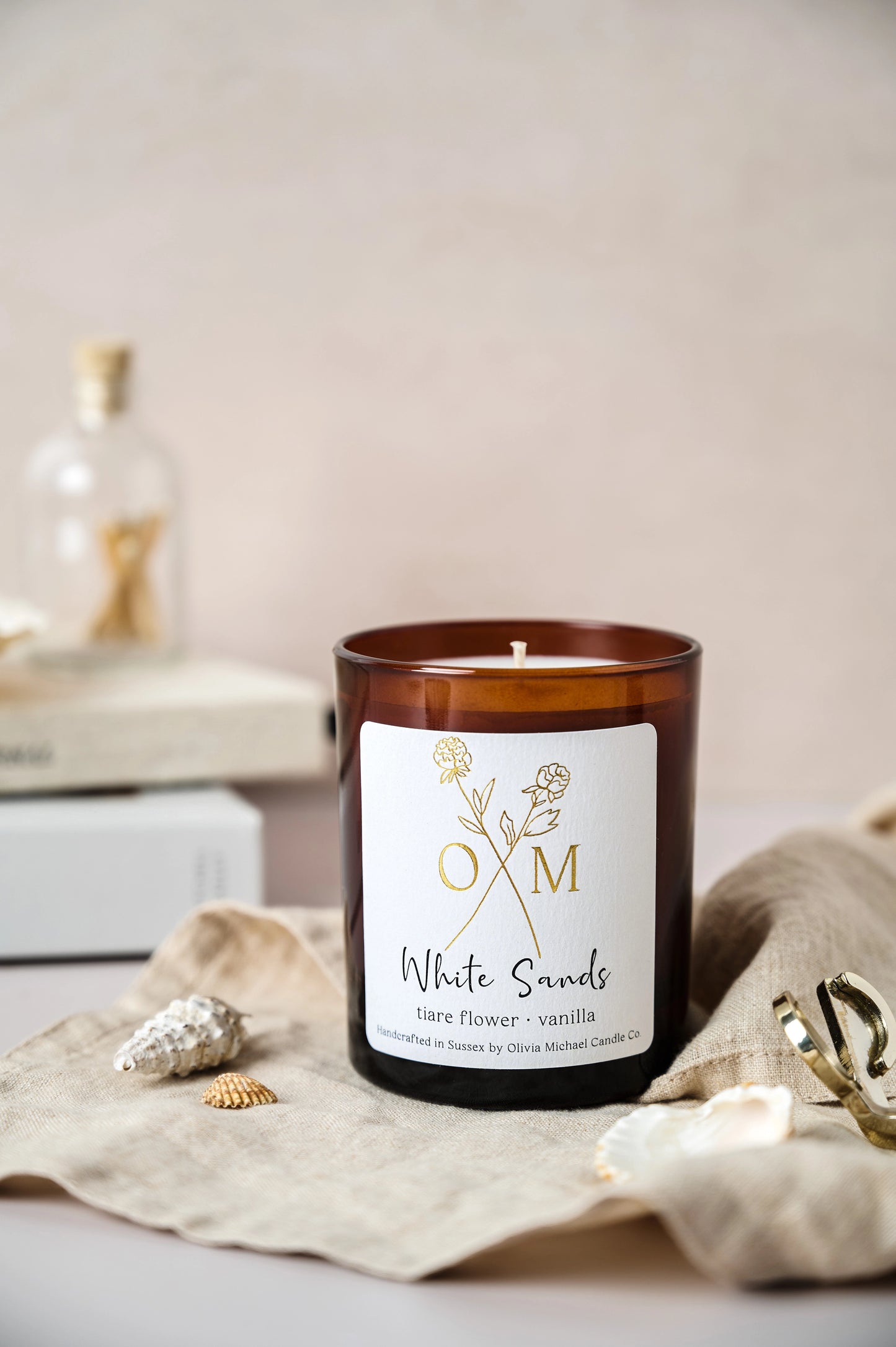 White Sands scented candle in a brown glass jar placed on a light beige cloth with small seashells and decorative items around it. The label reads 'White Sands, tiare flower and vanilla'.