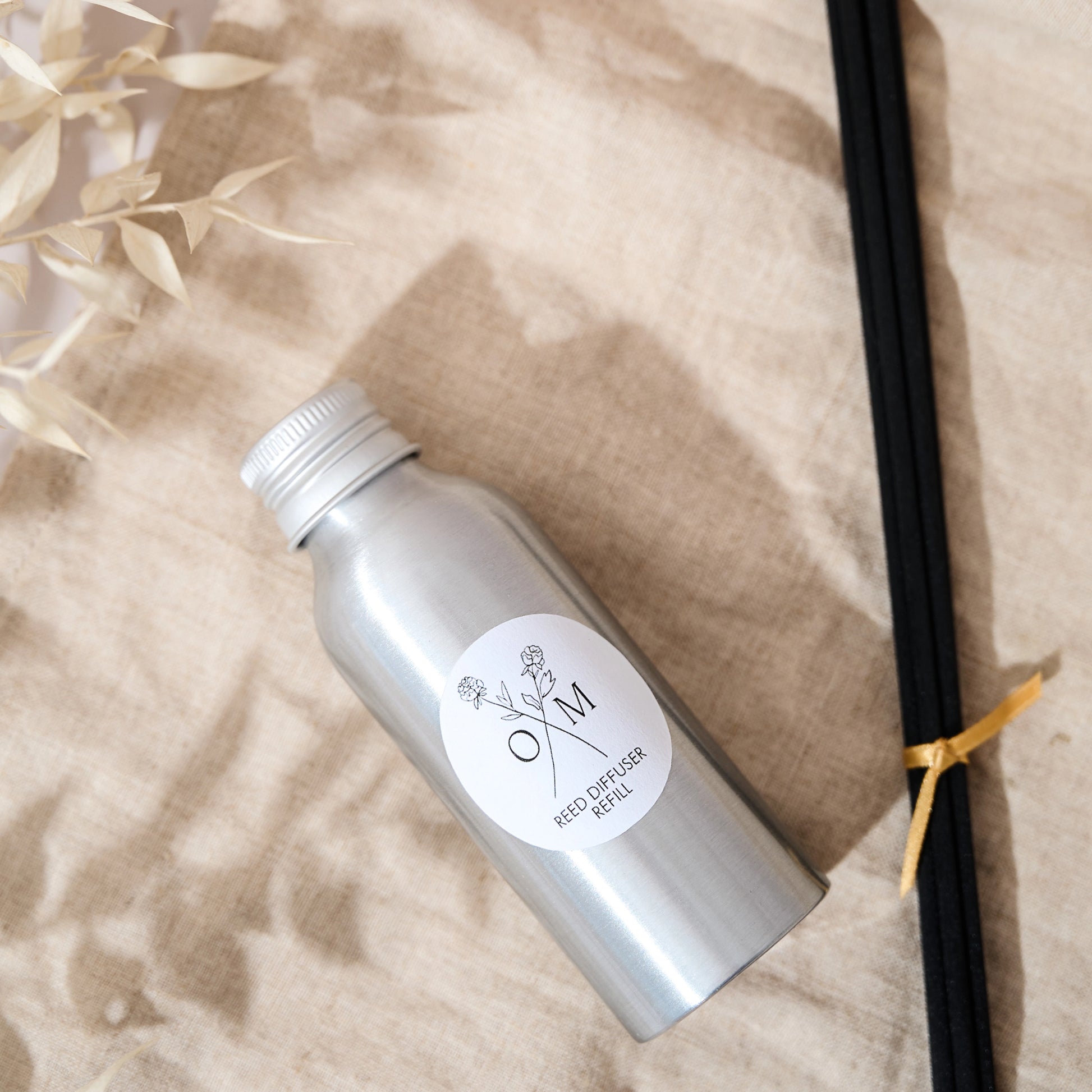 Reed diffuser refill in a silver bottle labeled 'Reed Diffuser Refill' placed on a light beige fabric next to a bundle of black reeds tied with a yellow ribbon.