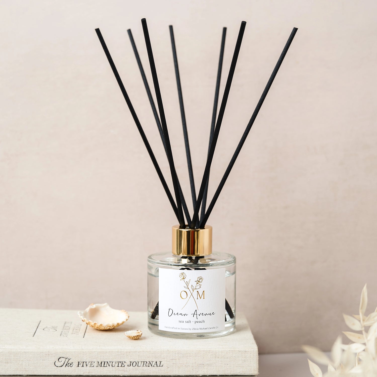 Ocean Avenue reed diffuser with black reeds and a gold cap, placed on top of 'The Five Minute Journal' with small seashells around it. The label reads 'Ocean Avenue, sea salt and peach'