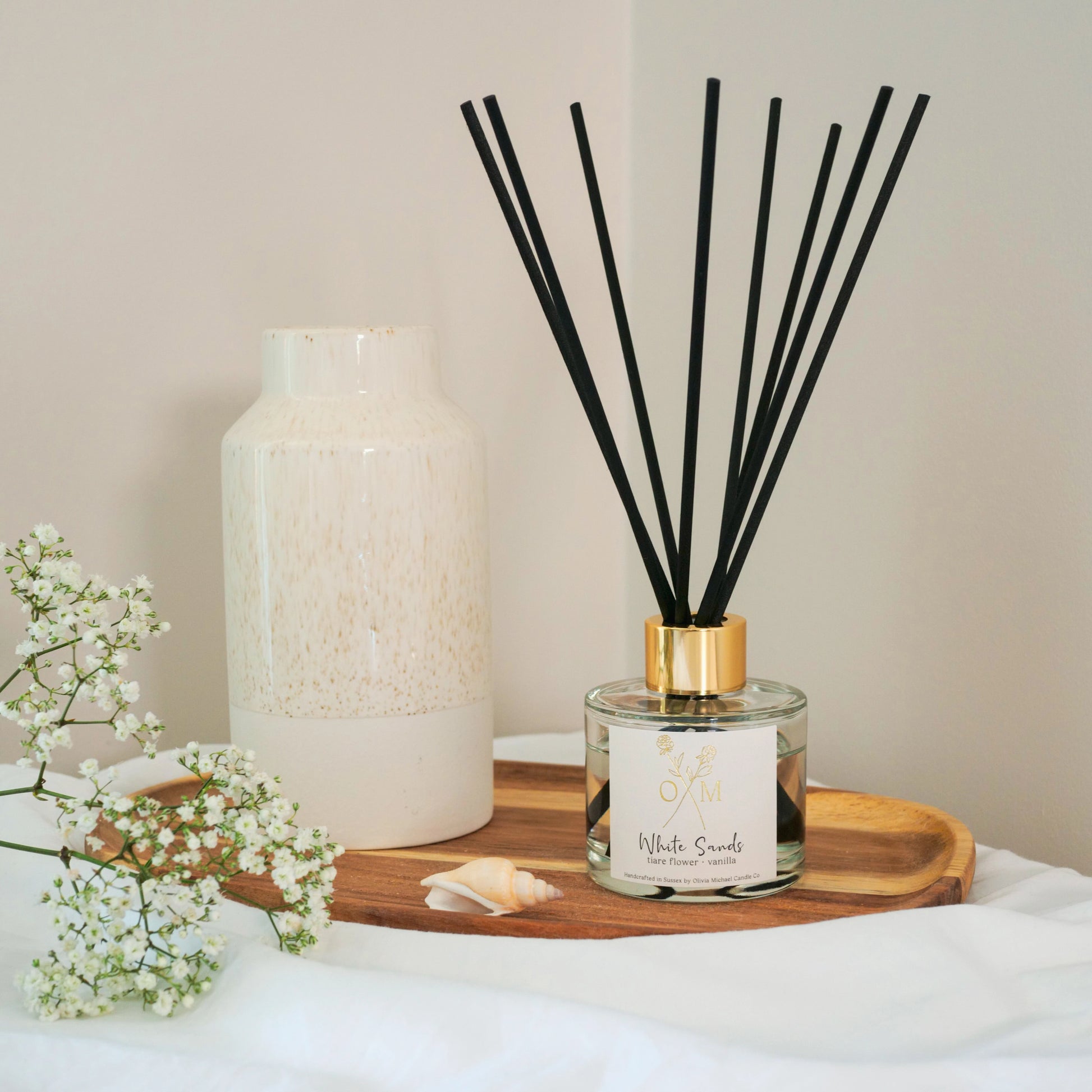 Elegant White Sands reed diffuser by Olivia Michael Candle Co. with black reeds, a gold-accented cap, and a tiare flower and vanilla fragrance, styled on a wooden tray beside a ceramic vase and delicate white flowers.