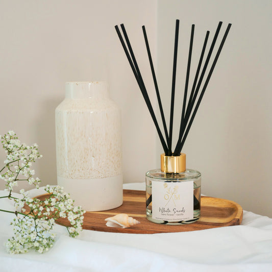 Elegant White Sands reed diffuser by Olivia Michael Candle Co. with black reeds, a gold-accented cap, and a tiare flower and vanilla fragrance, styled on a wooden tray beside a ceramic vase and delicate white flowers.