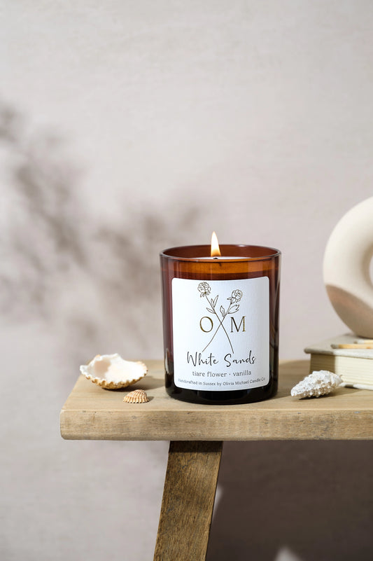 White Sands scented candle in a brown glass jar with a lit wick, placed on a wooden surface next to small seashells and decorative items. The label reads 'White Sands, tiare flower and vanilla'.