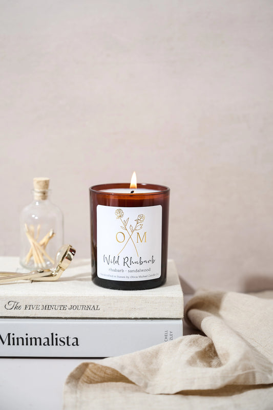 Wild Rhubarb scented candle in a brown glass jar with a lit wick, placed on top of 'The Five Minute Journal' and 'Minimalista' books. Next to the candle is a small glass jar with matches and a candle wick trimmer. The label reads 'Wild Rhubarb, rhubarb and sandalwood'.