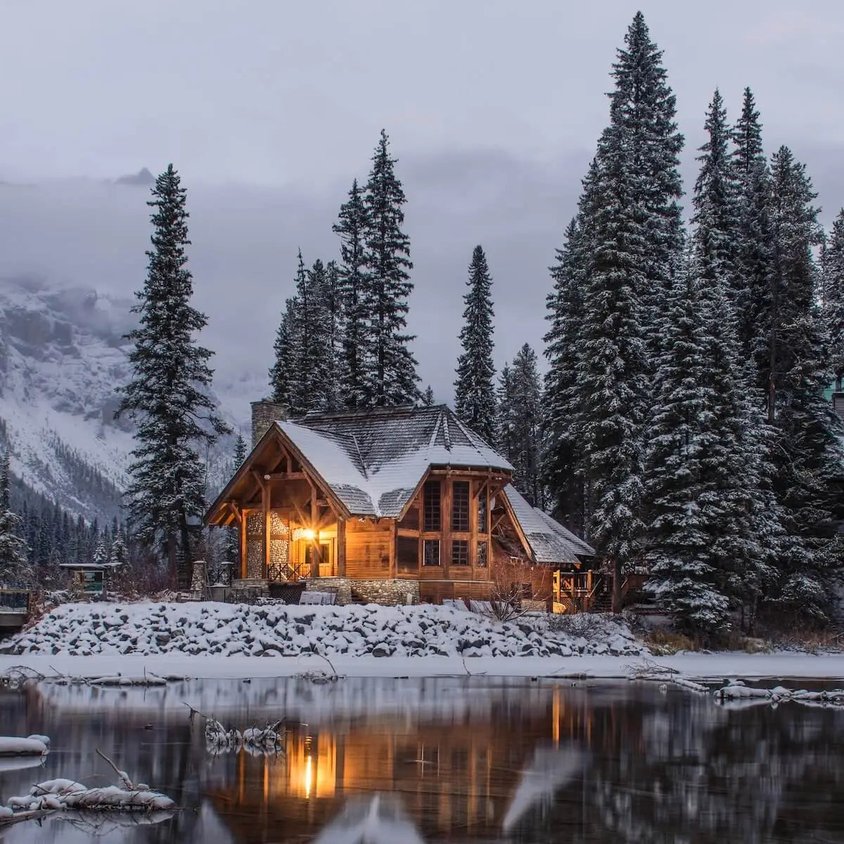 A cosy log cabin in amongst snowy mountains, a lake and snow tipped fir trees.